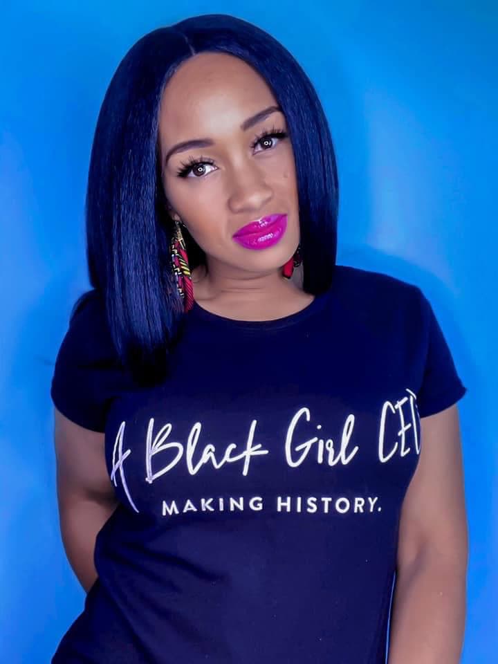 Hayley Surya Johnson headshot photo, woman with dark hair wearing pink lipstick a black tshirt with the words a black girl ceo making history on it standing in front of blue background
