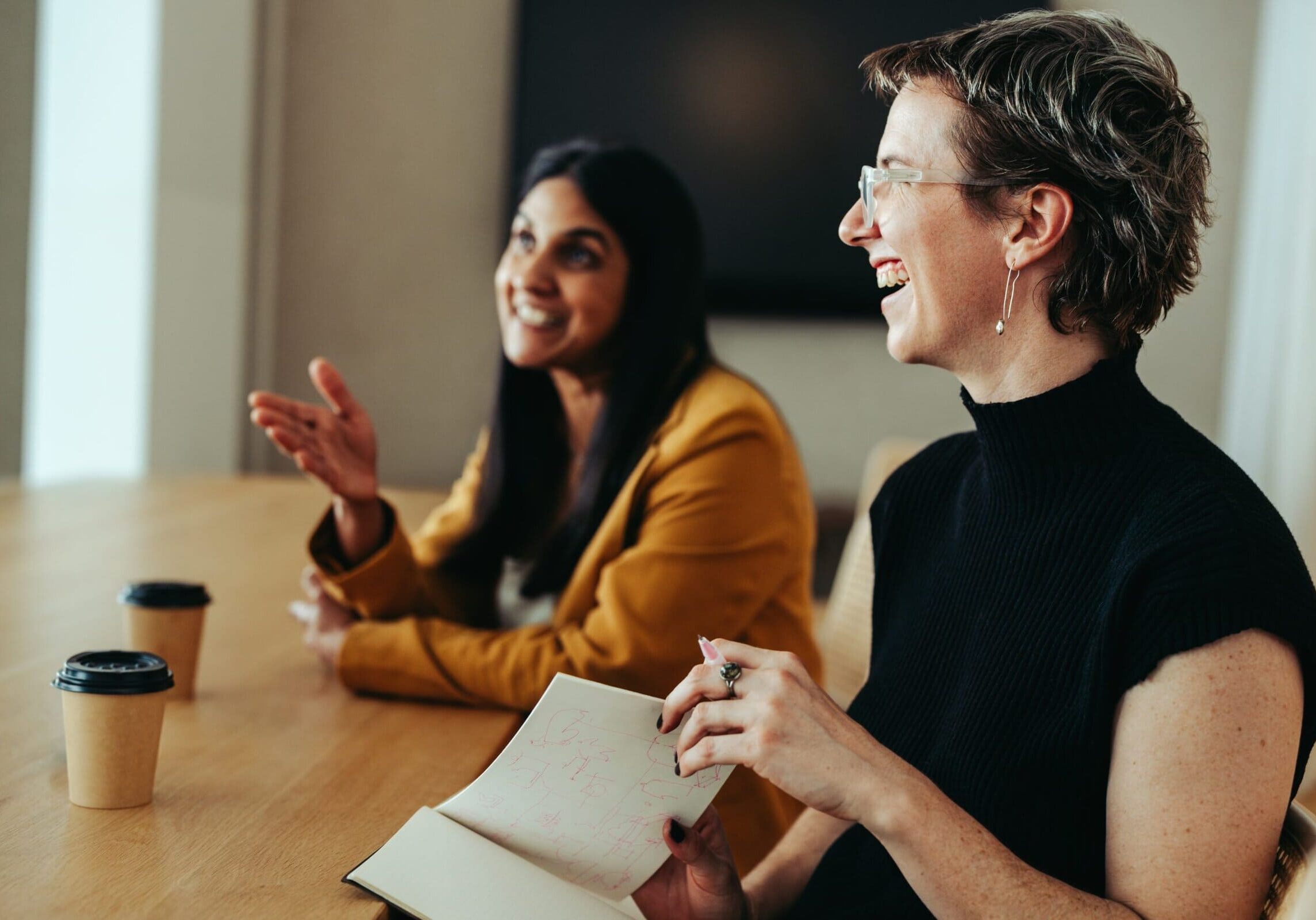 Two women smiling and talking while sitting at a table during a casual office meeting