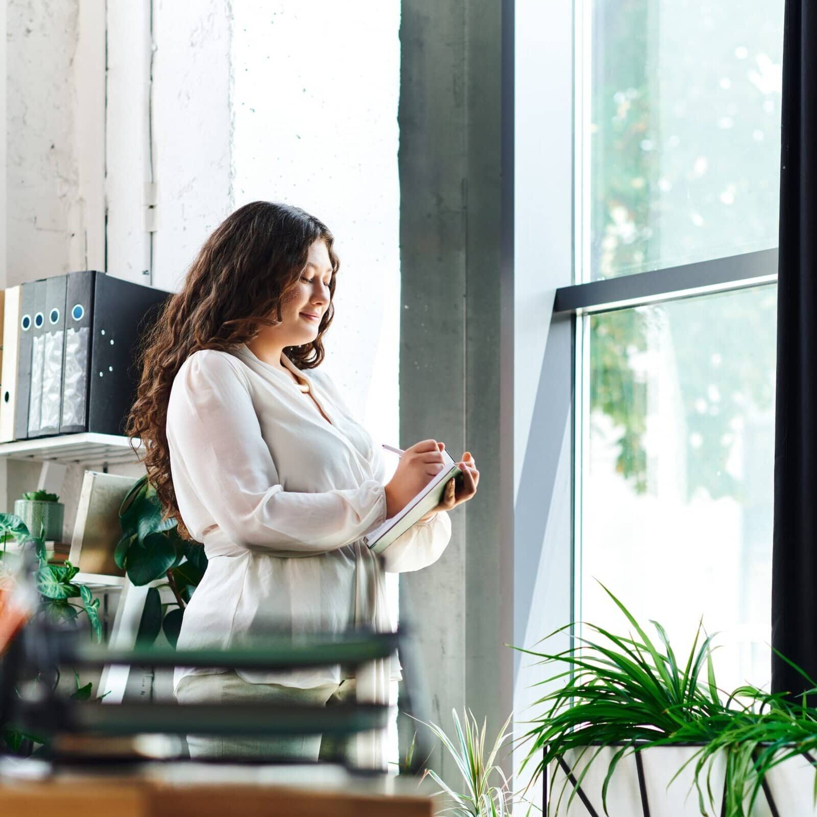 Confident woman standing by a window writing in a notebook in a bright office space