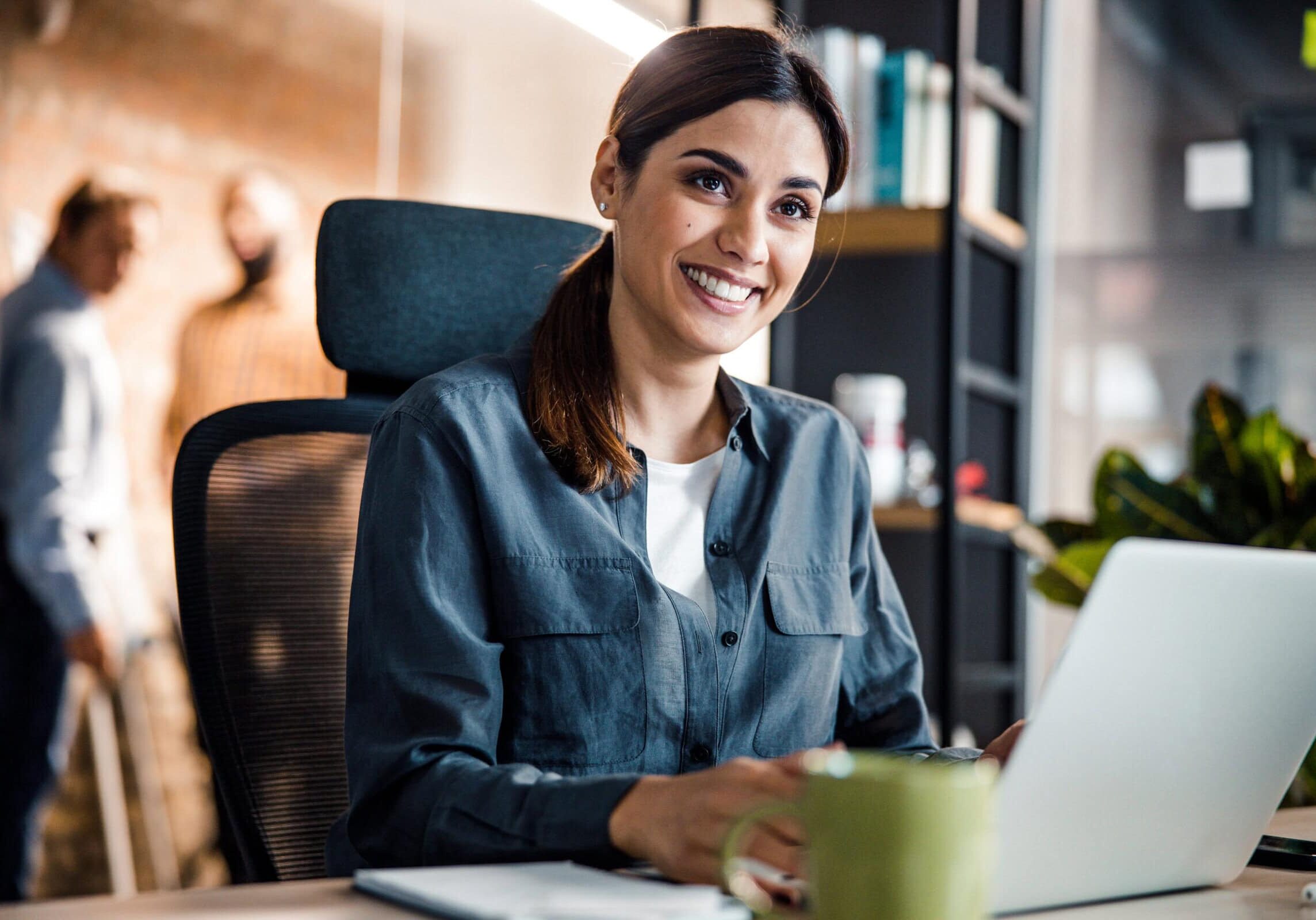 Woman smiling while working on a laptop at her desk in a modern offic