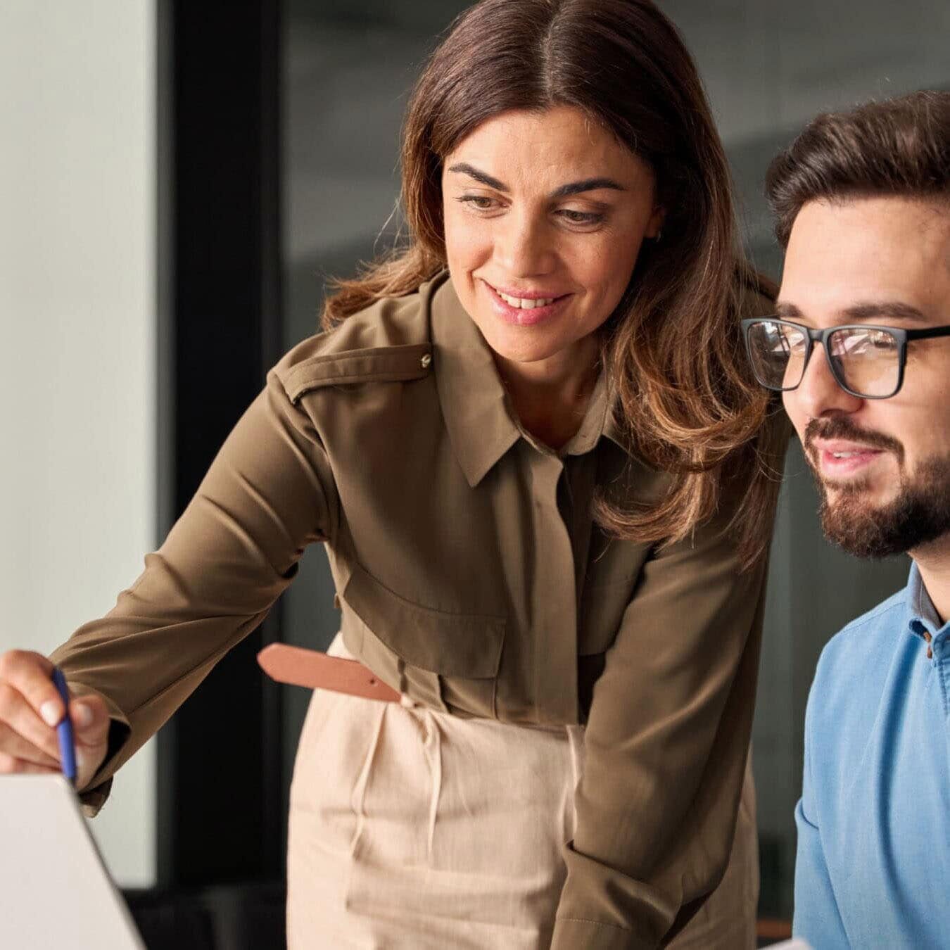 A woman helps a man on a laptop in an office setting