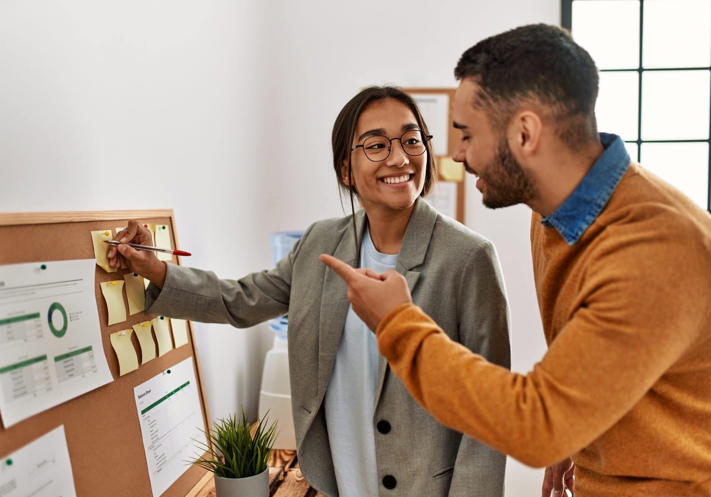 Two professionals discussing notes and charts on a corkboard in an office