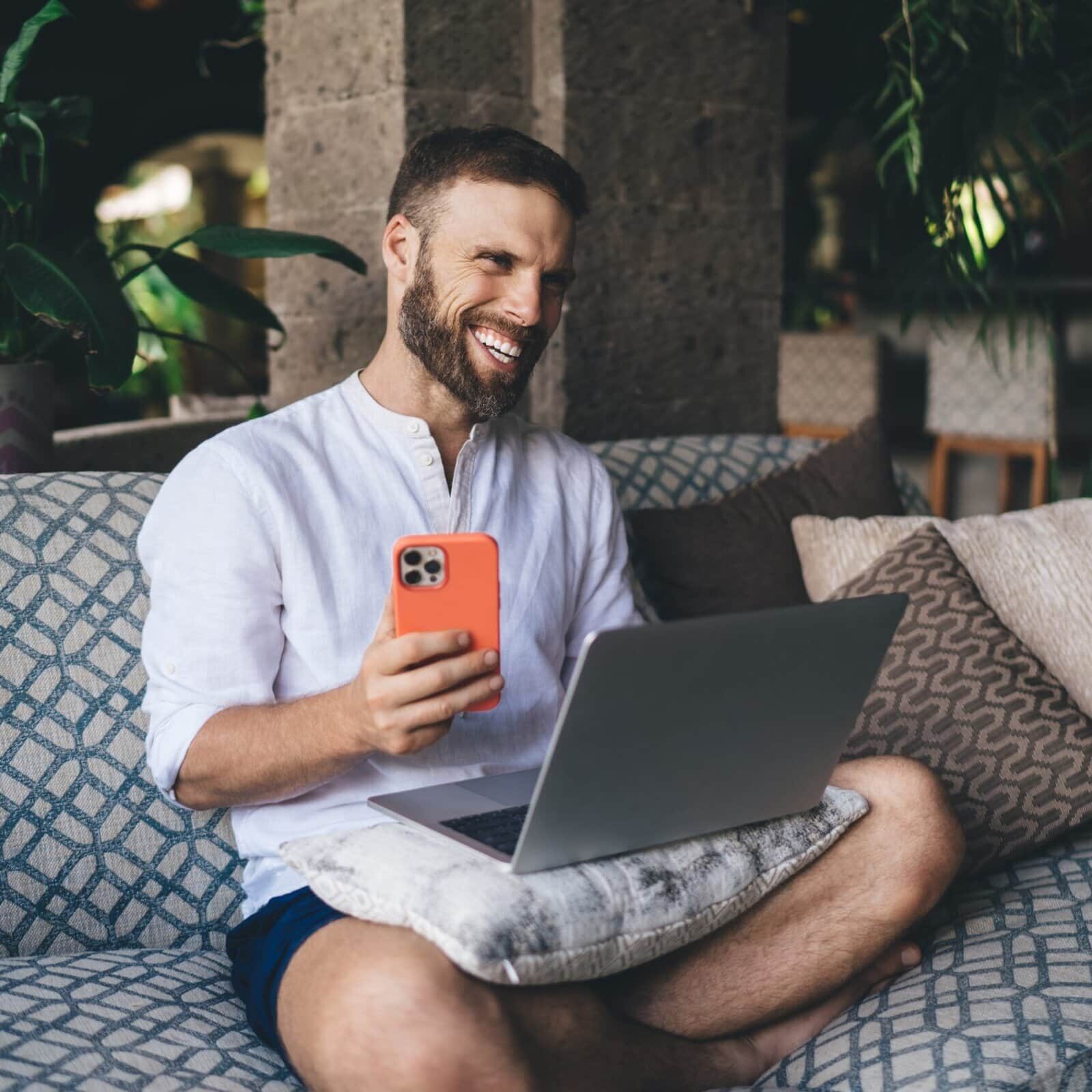 Man smiling while using a laptop and smartphone on a couch