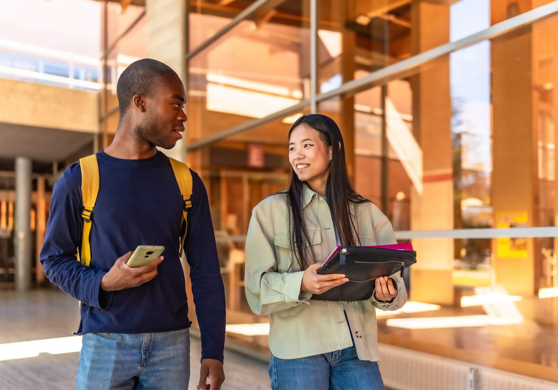 Two university students walking together outside a modern campus building, holding notebooks and talking