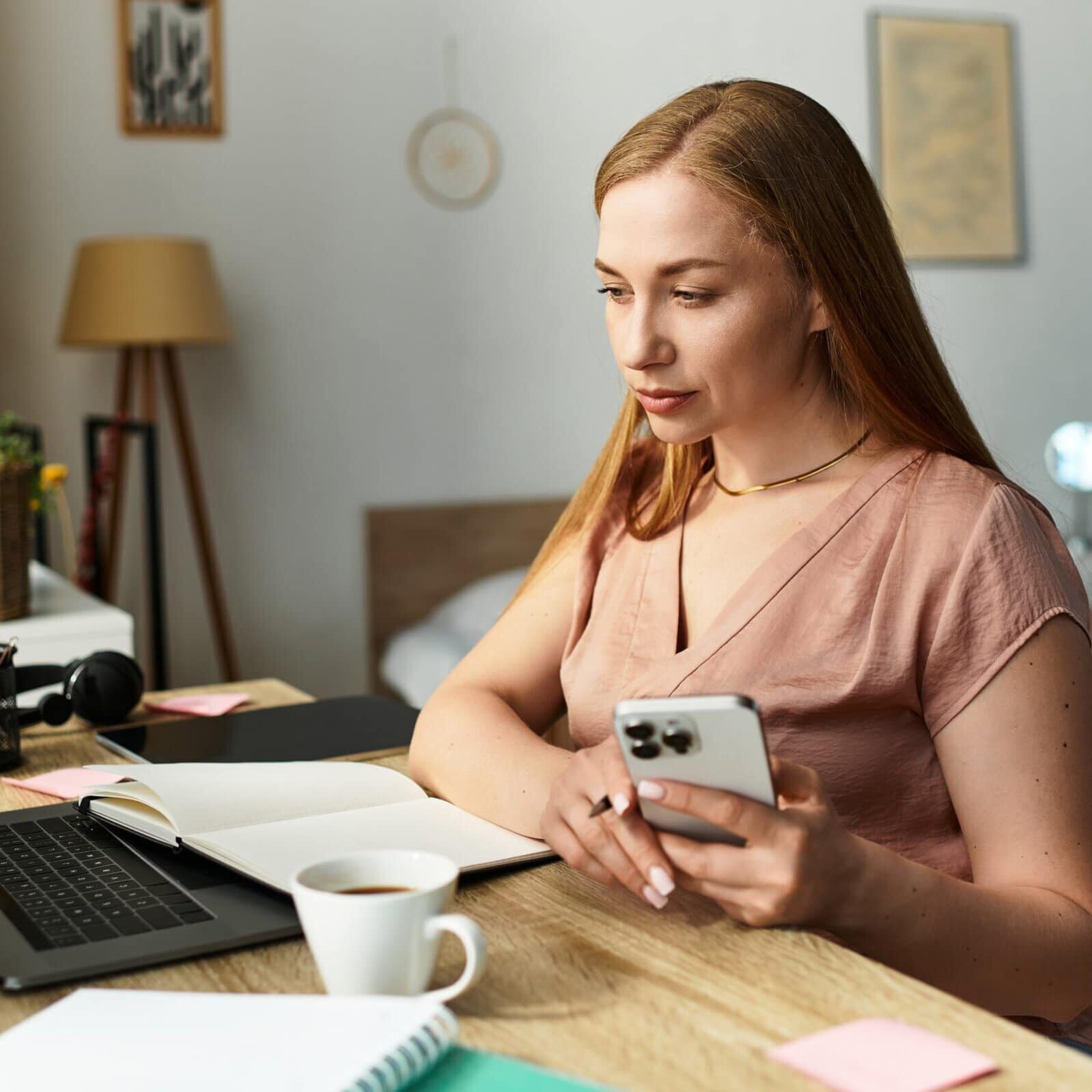Young woman sitting at a desk using a smartphone beside an open laptop and notebook