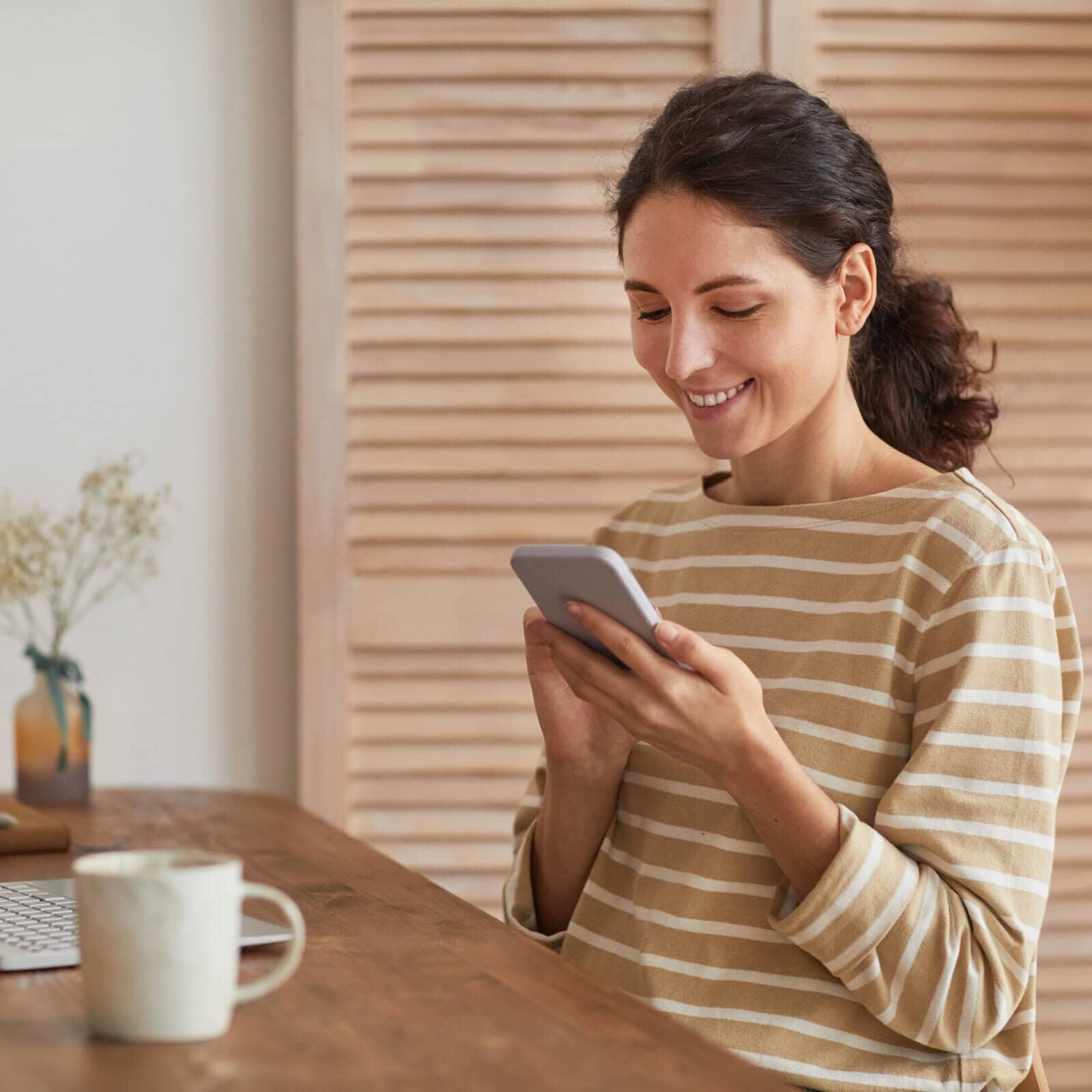 Woman smiling while using her smartphone at a home workspace with a laptop nearby.