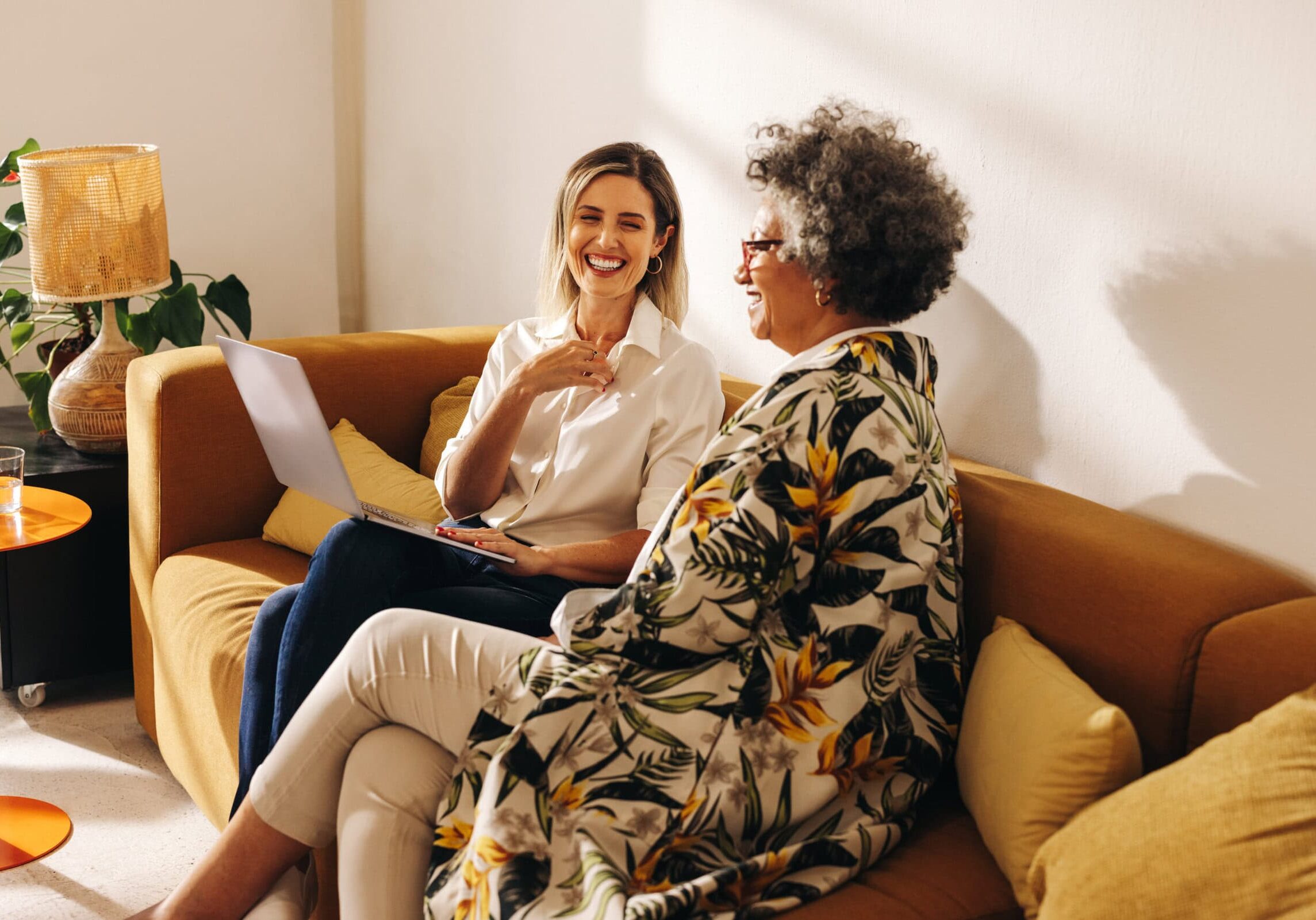 Two women engaged in a friendly coaching session on a couch, with one holding a laptop, in a cozy, sunlit room.