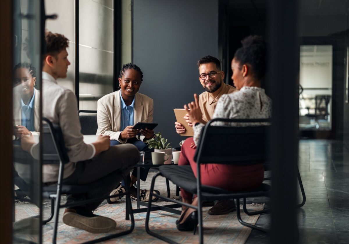 A diverse team of four professionals of different ethnic backgrounds is seated in a relaxed, modern office setting. They are engaged in a collaborative discussion around a small coffee table. One woman holds a tablet, another man holds a notebook, and the group appears focused and supportive, embodying the concept of workplace allyship and teamwork.