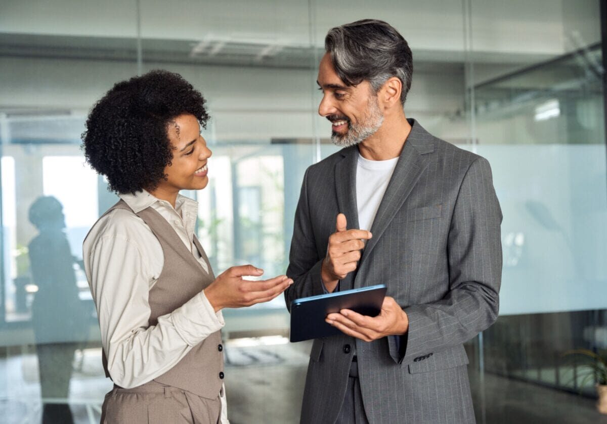 Two diverse professionals discuss ideas while standing together, with one holding a tablet during a coaching-style conversation.