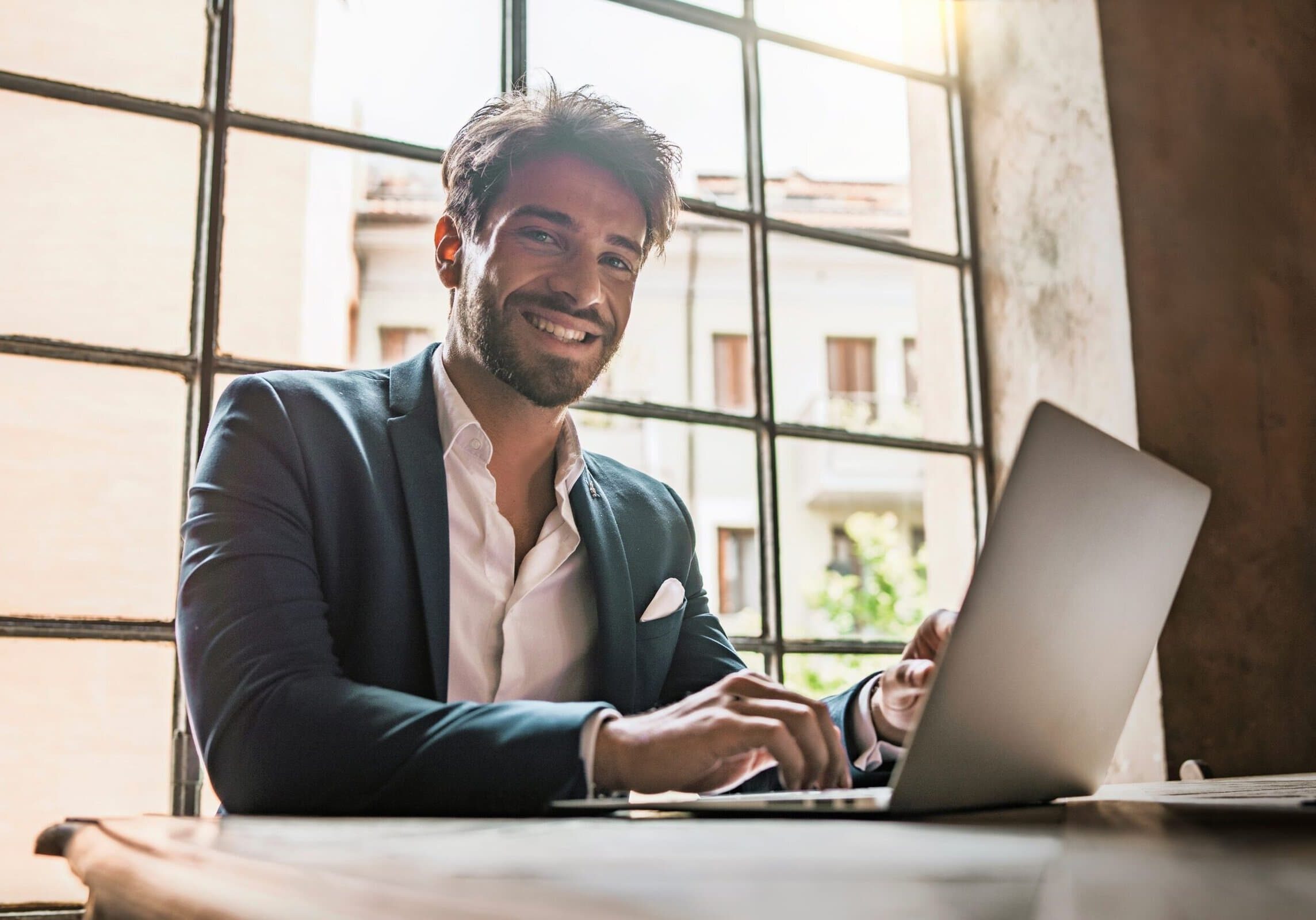 Smiling businessman working on a laptop at a desk near a large window in an office
