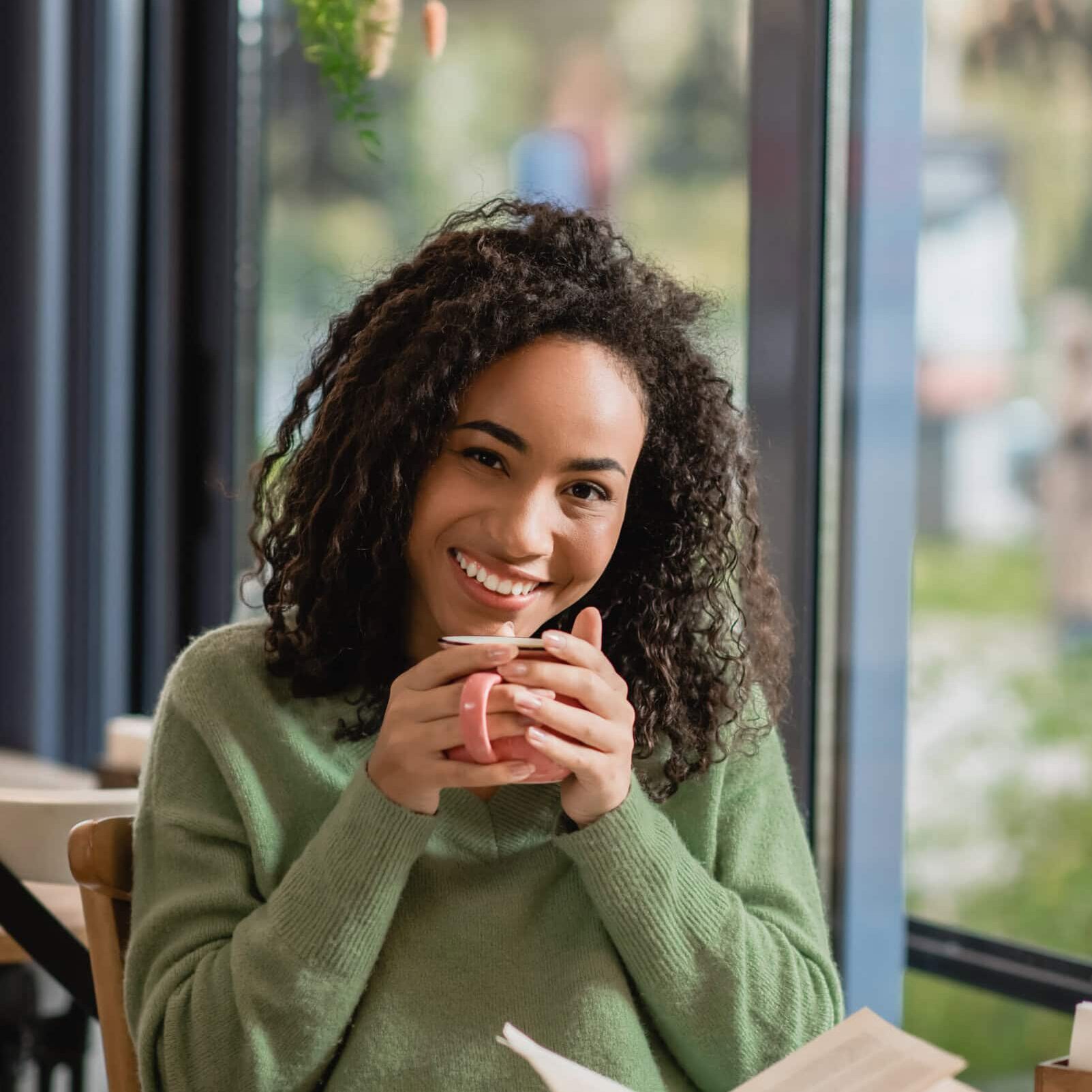 Smiling woman holding a cup of coffee while reading a book at a table by a window