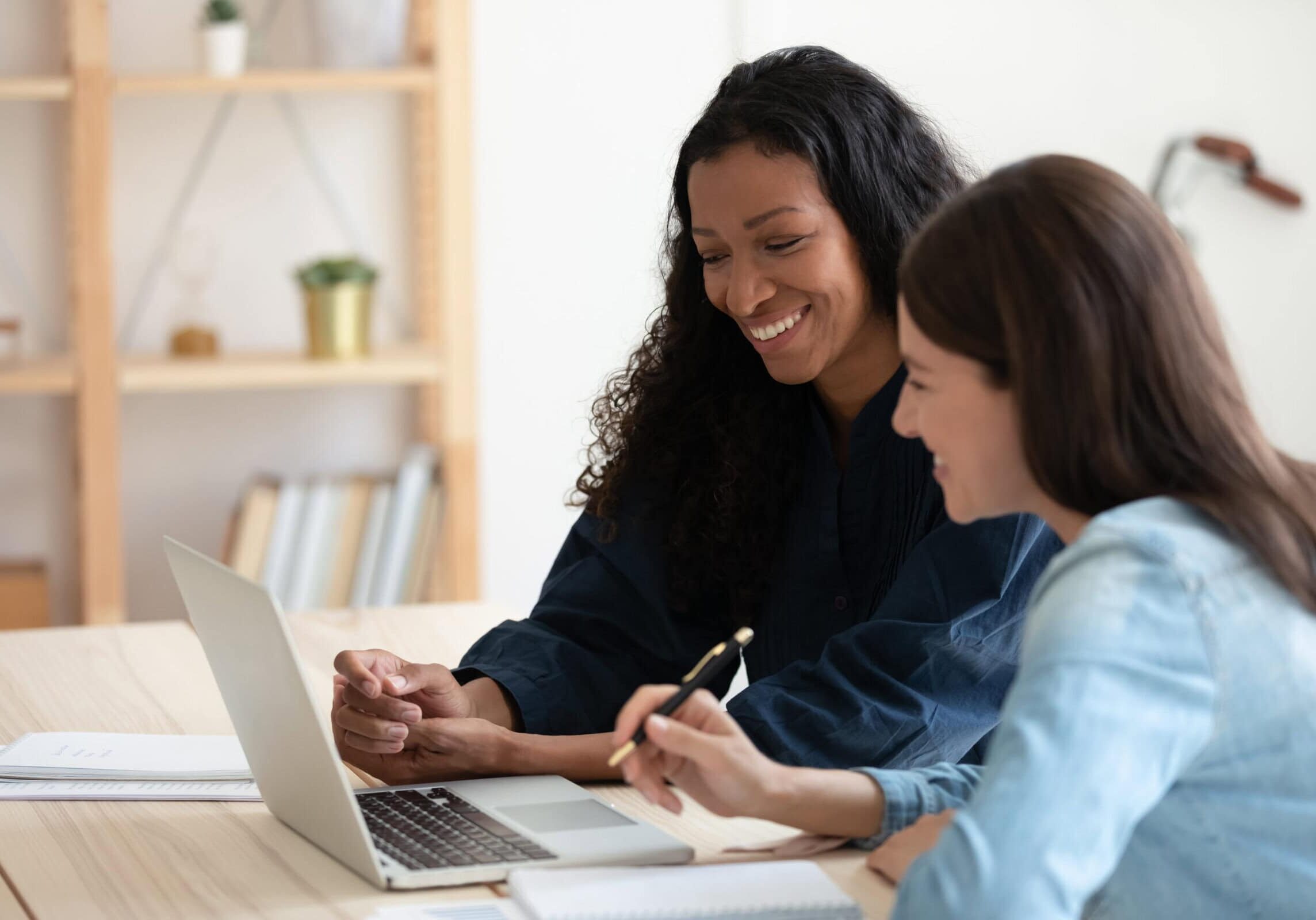 Two women smiling while reviewing information together on a laptop at a desk