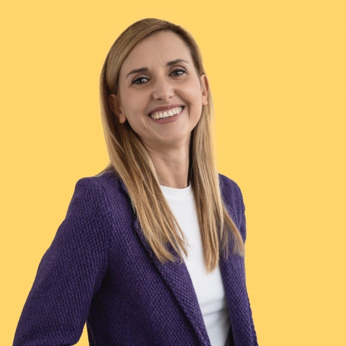 Elena Giorgetti PCC headshot photo, smiling woman with blond hair wearing purple blazer over a white shirt in front of yellow background