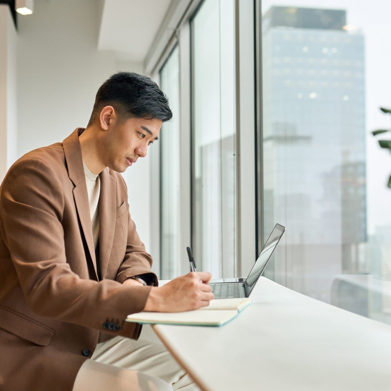 man sitting at table in front of window taking notes in a notebook while looking at a laptop