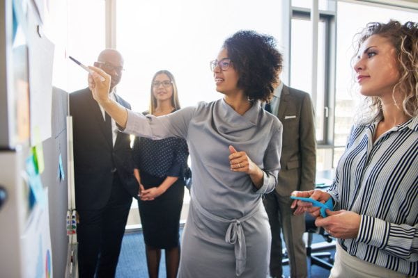 coach pointing to something on a whiteboard with four others listening, all standing in a conference room