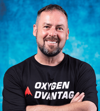 Patrick McKwoen headshot photo, smiling man wearing black long sleeve shirt with logo Oxygen Advantage in front of blue background